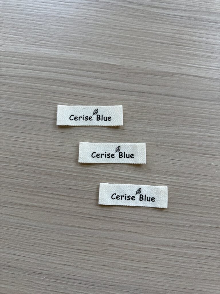 Three light-coloured woven-label samples with dark CeriseBlue lettering and a small leaf motif